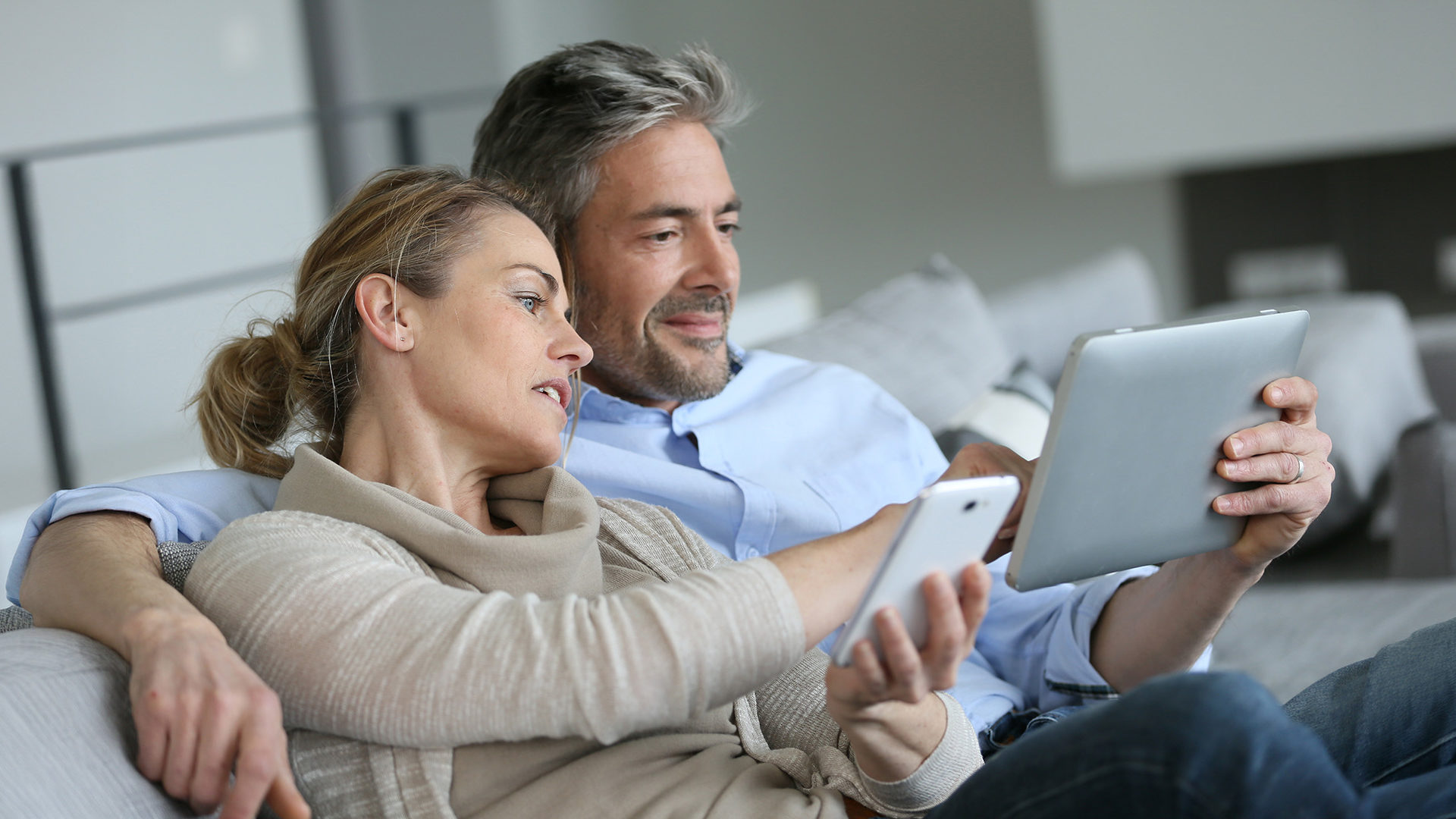 Couple sitting on sofa researching on their tablet and mobile device.