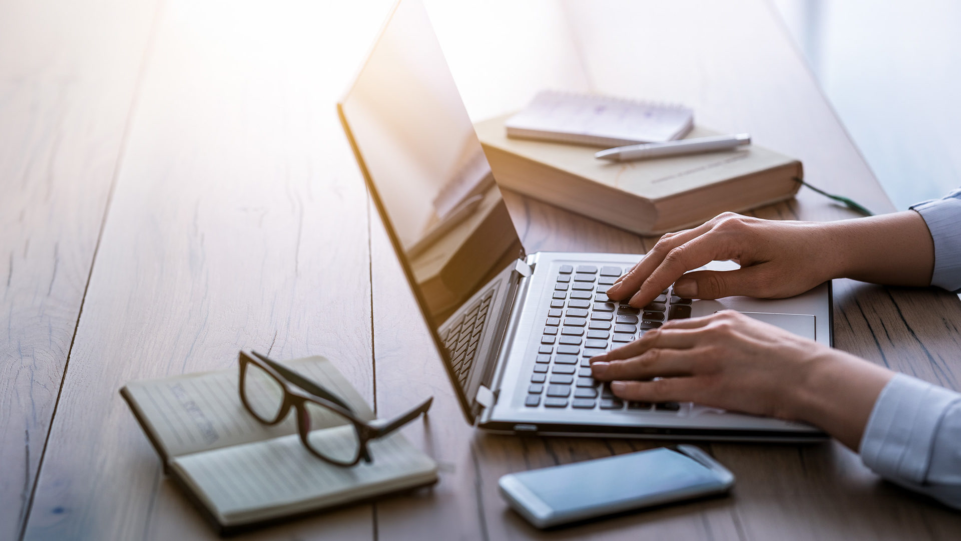 Hands on a laptop keyboard with glasses and books on a desk.