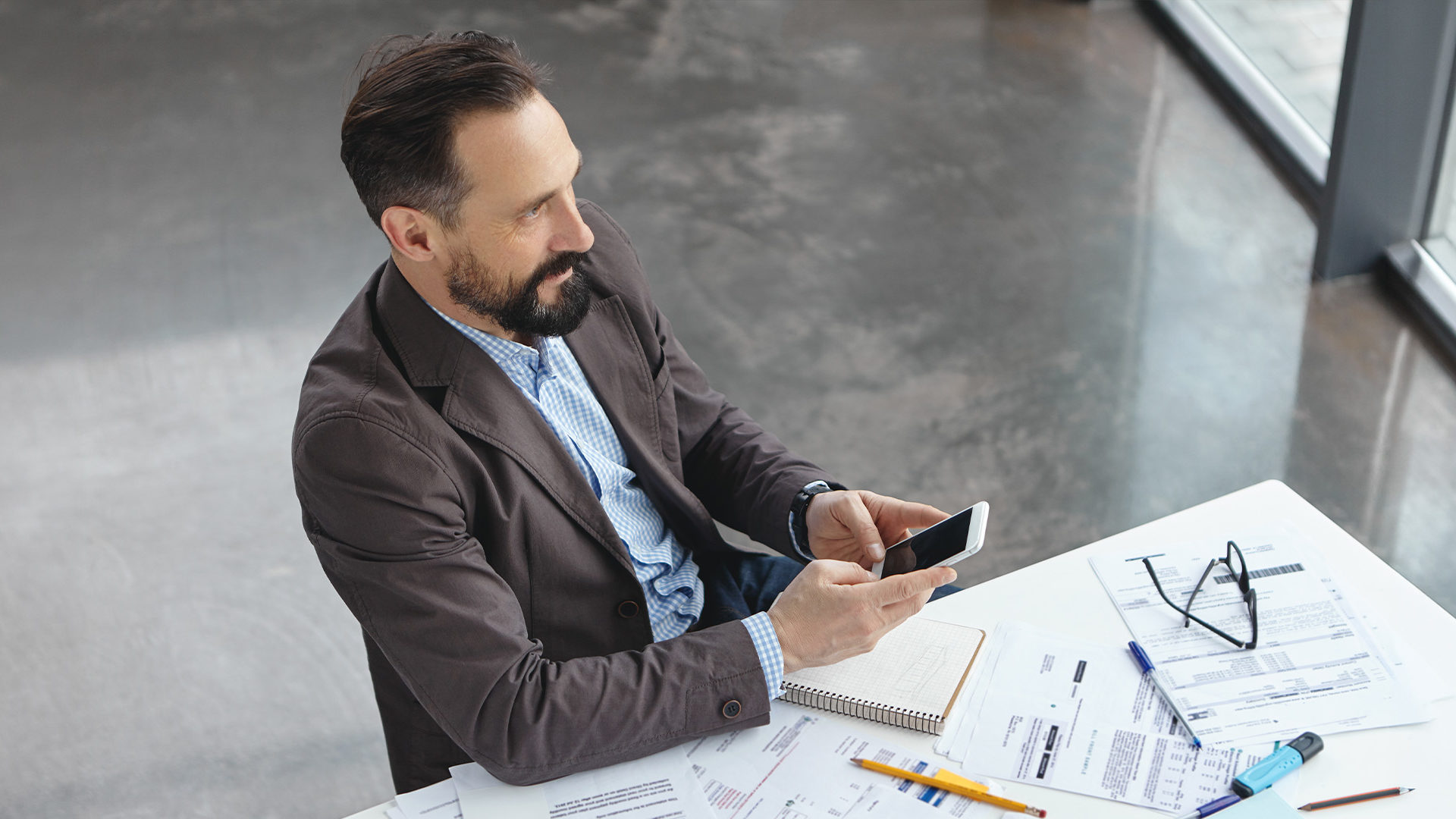 Business owner sitting at desk with paperwork and looking at mobile device.