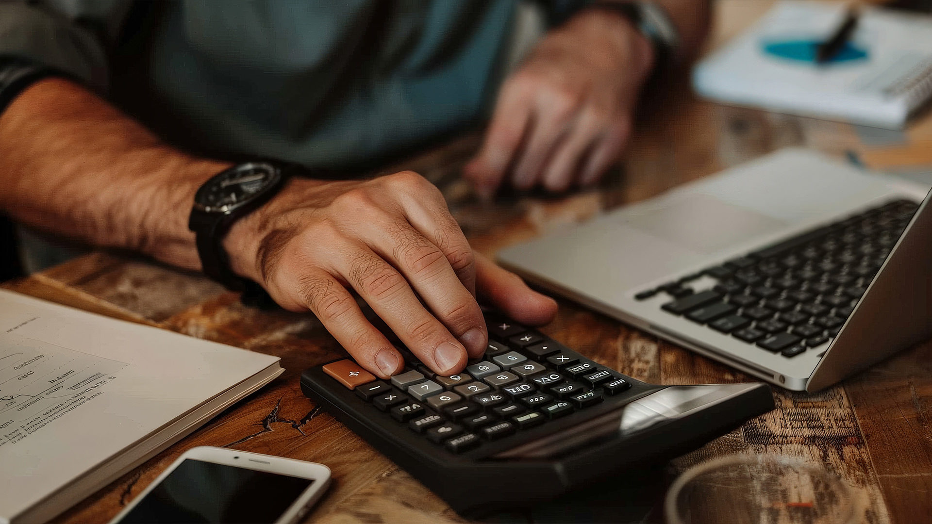 Hand pressing buttons on a calculator with a laptop, mobile phone and paperwork at a desk.