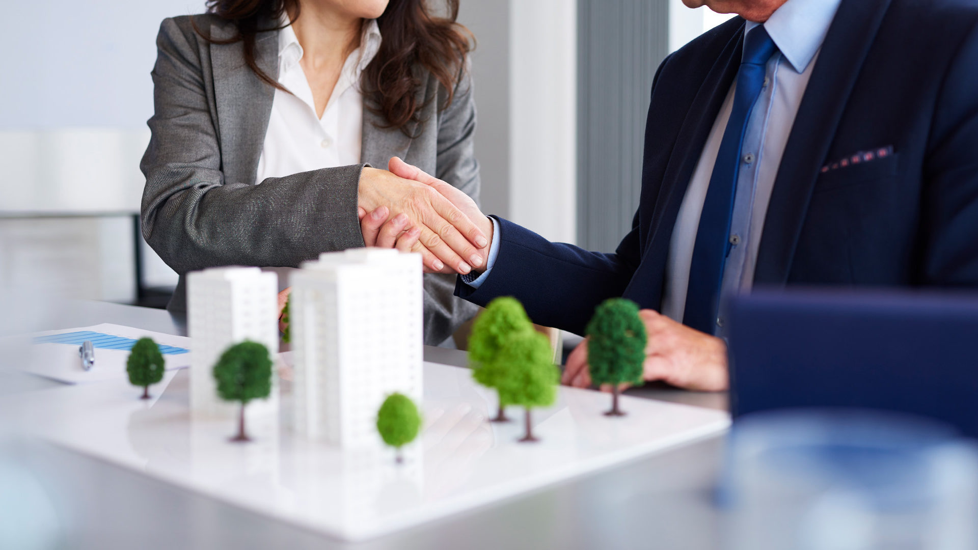 Two business people shaking hands with model building and trees on the table.